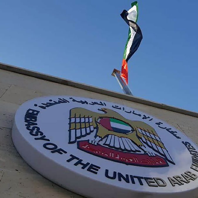 تظهر في الصورة شعار سفارة الإمارات العربية المتحدة. الشعار يتواجد على جدار السفارة، ويعلوه علم الإمارات الذي يرفرف في السماء. الخلفية تشمل السماء الزرقاء مع بعض السحب البيضاء. التصميم يعكس الهوية الوطنية ويظهر الفخر بالبلد.