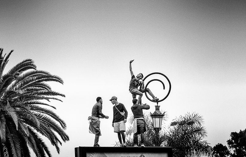 L'image montre un groupe de personnes en action, probablement en train de réaliser une performance acrobatique ou un numéro de cirque. L'une des personnes est perchée au sommet d'un cercle, tandis que les autres l'entourent, soutenant ou aidant dans la mise en scène. Le fond est dominé par un ciel clair et des palmiers, ce qui donne une ambiance estivale à la scène. L'ensemble est photographié en noir et blanc, ajoutant une touche artistique et dramatique.