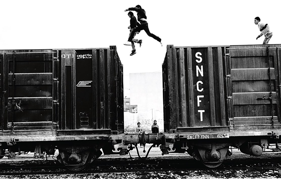 L'image montre un groupe de jeunes qui sautent d'un wagon de train à un autre. La scène est en noir et blanc, ce qui accentue l'action dynamique et l'intensité du moment. On peut observer deux jeunes sautant dans les airs, tandis que d'autres se trouvent sur les wagons ou à proximité. L'environnement semble urbaine, avec une atmosphère à la fois aventureuse et un peu risquée. Les détails des wagons et l'arrière-plan renforcent l'impression d'un cadre industriel.