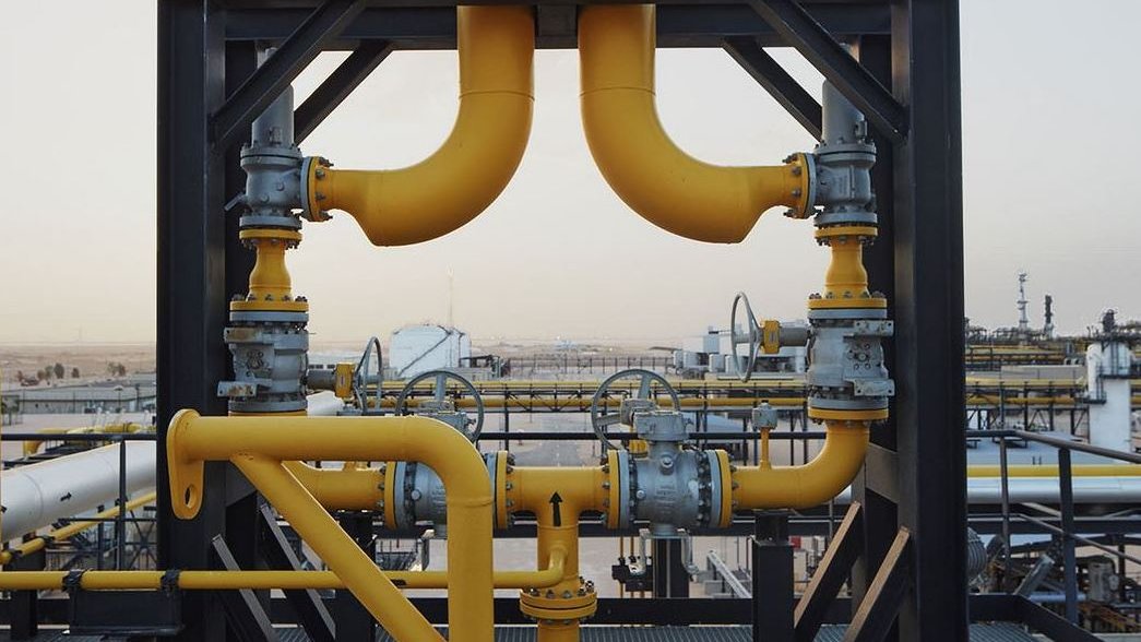 L'image montre un réseau de tuyauterie industrielle. On peut voir des tuyaux en métal, peints en jaune, qui se croisent et se connectent à différentes valves et équipements. L'arrière-plan présente un paysage industriel, avec d'autres structures et installations visibles au loin, suggérant un environnement de production ou de traitement de ressources. Le tout semble bien organisé et conçu pour la circulation de fluides, probablement dans le secteur pétrolier ou gazier.