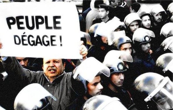 L'image montre une scène de manifestation où un homme tient une grande pancarte avec l'inscription "PEUPLE DÉGAGE !" Au premier plan, cet homme semble déterminé et engagé, tandis qu'en arrière-plan, un groupe de policiers en uniforme avec des casques se tient à proximité, suggérant une tension entre les manifestants et les forces de l'ordre. L'atmosphère semble chargée d'émotion et de revendication.