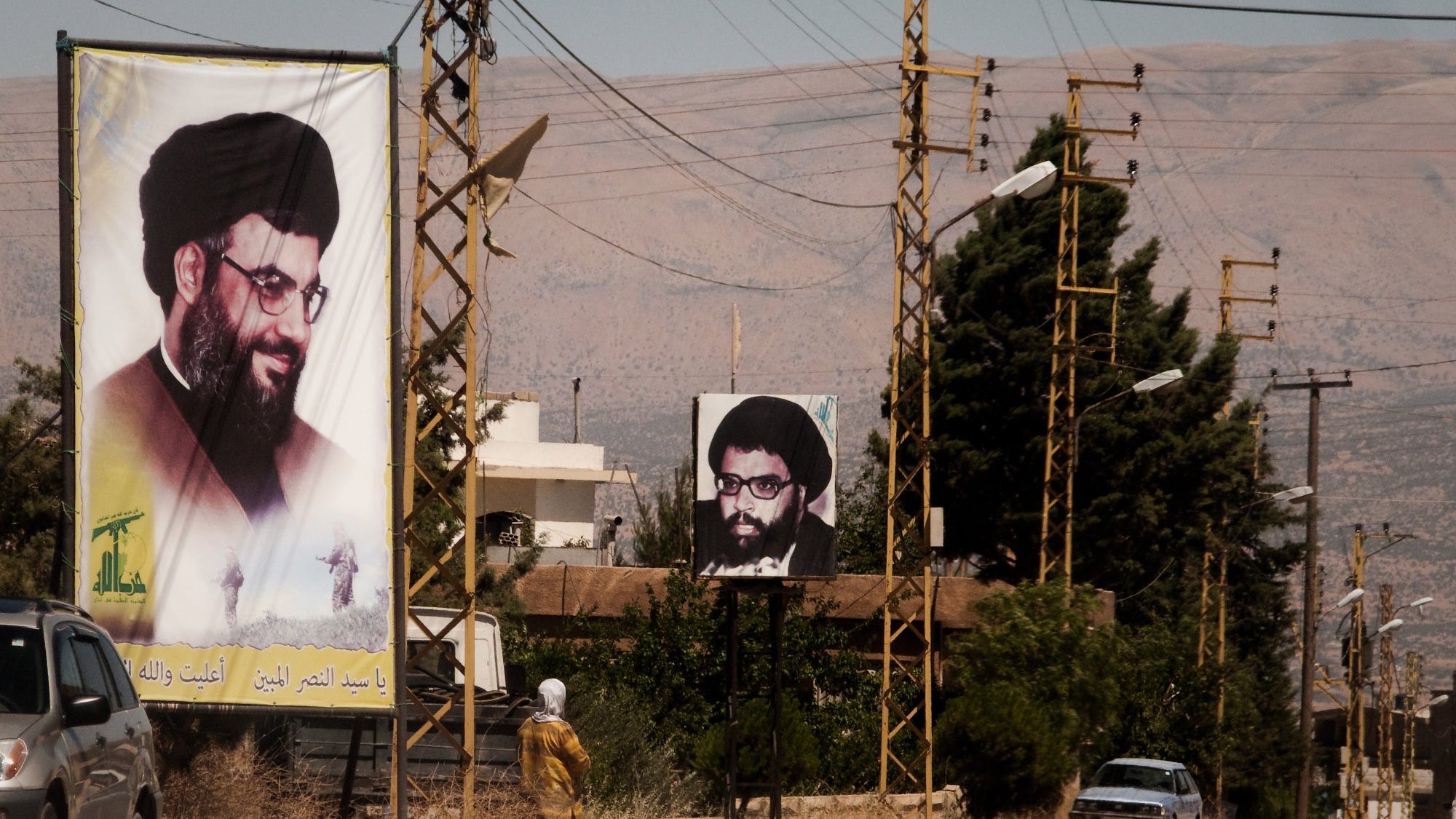 L'image montre une rue avec des panneaux d'affichage sur lesquels figurent des portraits d'un homme portant des lunettes et une barbe, associé à une ambiance rurale. On peut également voir des lignes électriques et quelques bâtiments en arrière-plan. À gauche, un panneau est en couleur et à droite, un autre est en noir et blanc. La scène semble être située dans une région montagneuse. C'est une illustration d'un espace public où des affiches politiques ou religieuses peuvent être observées.