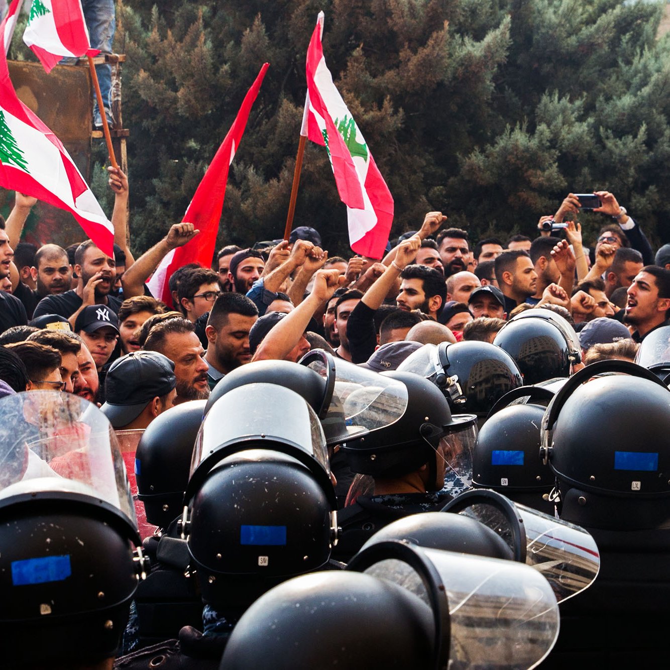 L'image montre une scène de manifestation avec un grand nombre de personnes brandissant des drapeaux libanais. Les manifestants semblent exprimer leurs revendications avec des poings levés, tandis qu'une ligne de policiers en uniforme, équipés de casques et de protections, se tient face à eux. L'ambiance est tense, illustrée par la proximité des manifestants et des forces de l'ordre. Des arbres en arrière-plan complètent le décor de cette confrontation.