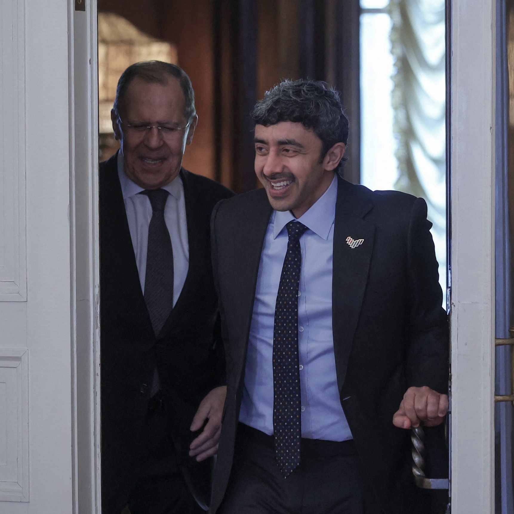 The image shows two men entering a room together. One man is smiling and appears to be in a suit, indicating a formal occasion. The other man, who seems more serious, is walking closely behind. The setting looks elegant, likely a governmental or official location, with decorative elements visible in the background. The overall mood suggests a friendly or cordial interaction between the two individuals.