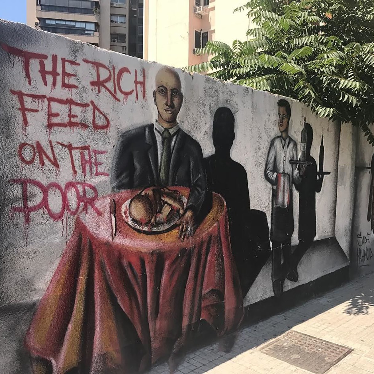 The image features a mural on a wall depicting two main figures. On the left, a man in a suit sits at a table covered with a red tablecloth, holding a loaf of bread and looking directly at the viewer. To the right, another figure dressed as a waiter stands with a tray, while dark shadows cast behind them suggest more figures or a hidden narrative. The phrase "THE RICH FEED ON THE POOR" is painted prominently nearby, conveying a theme of social inequality and critique of class disparity. The background includes urban elements like buildings and greenery, adding to the setting of the mural.