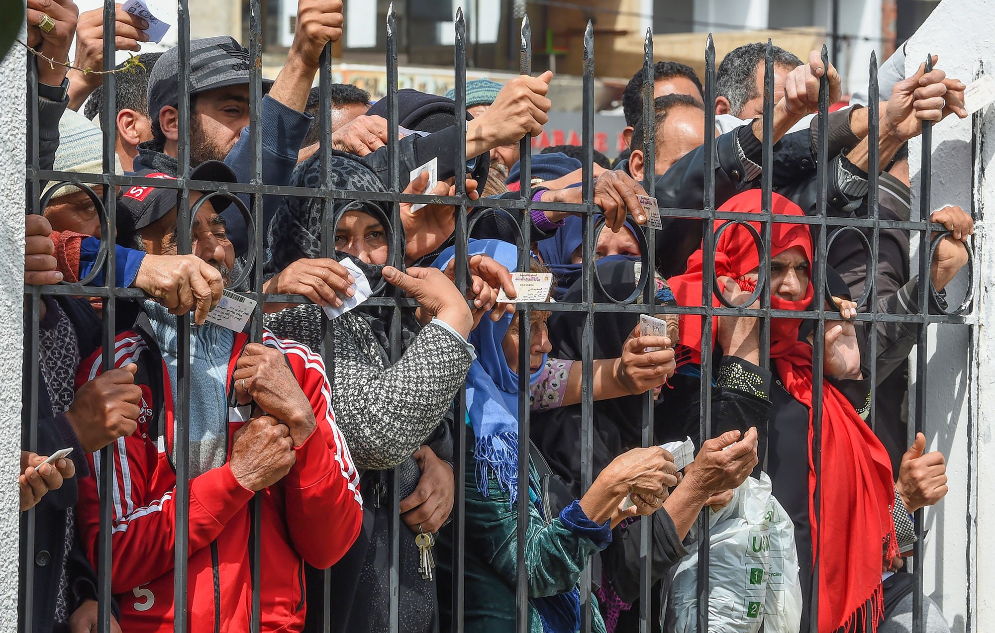 تظهر في الصورة مجموعة من الأشخاص المتجمعين خلف سياج حديدي. يرفع البعض منهم أوراقًا أو بطاقات، بينما يبدو التركيز على حركتهم وتصرفاتهم، مما يوحي بأنهم قد يكونون في انتظار شيء ما أو يحاولون الوصول إلى جهة معينة. الأجواء تعكس حالة من الترقب أو الأمل، مع تنوع في الملابس، مما يشير إلى خلفيات مختلفة.