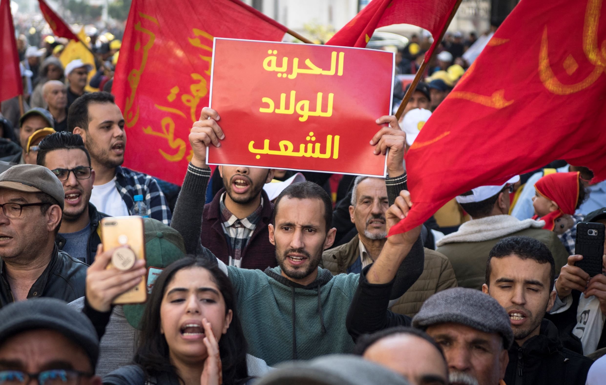 Nell'immagine si vede una manifestazione di protesta affollata, con persone che sventolano bandiere rosse. In primo piano, un giovane tiene in alto un cartello con scritto in arabo "libertà per il popolo". Altre persone intorno a lui sembrano partecipare attivamente, alcune con telefoni in mano per immortalare il momento. L'atmosfera è carica di emozione e determinazione, riflettendo il desiderio di cambiamento e di diritti.