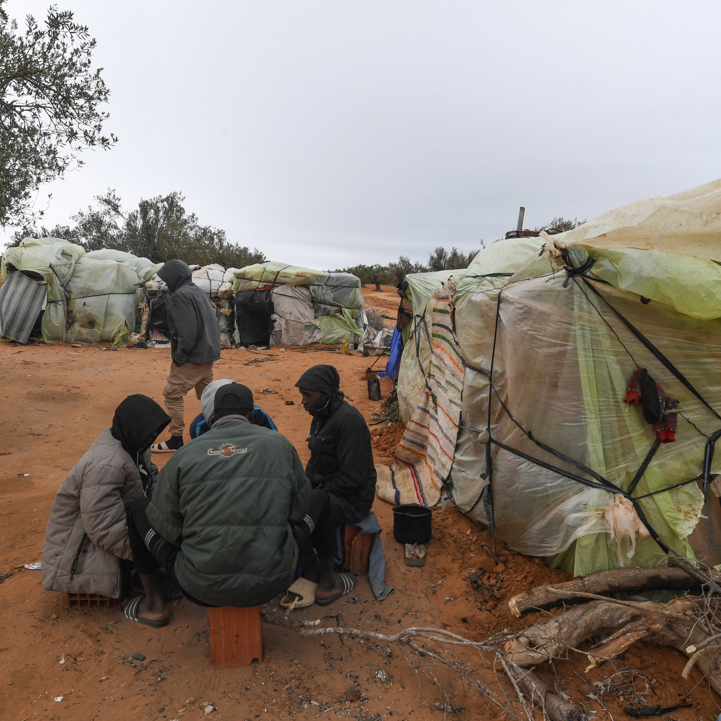 L'image montre un groupe de personnes assises ensemble sur le sol, entourées de tentes faites de matériaux recyclés, telles que des bâches en plastique. Le décor est désertique, avec un sol de sable et quelques arbres à l'arrière-plan. Les personnes semblent être en conversation ou en train de partager quelque chose. Le temps paraît nuageux, ajoutant une atmosphère sombre à la scène.