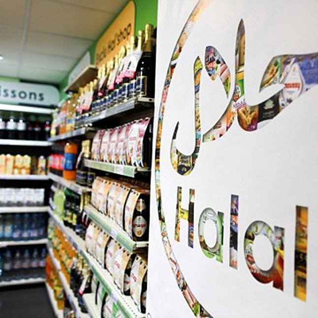 L'image montre une vue intérieure d'un magasin avec une étagère remplie de divers produits alimentaires et boissons. Au premier plan, il y a un grand logo représentant le mot "Halal" en lettres stylisées. Ce logo est intégré dans un design créatif qui semble inclure des images de produits alimentaires. L'éclairage est clair, et l'ambiance générale de la scène est celle d'un espace de vente organisé et vibrant.