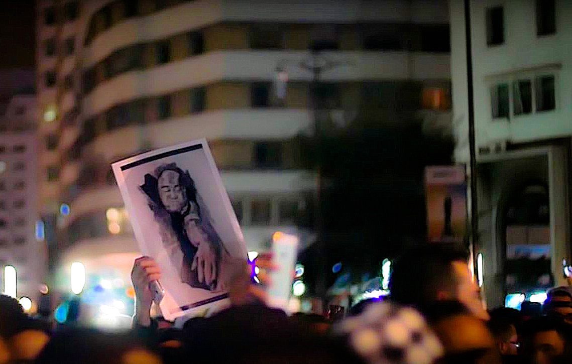 تظهر الصورة مجموعة من الأشخاص في مظاهرة أو تجمع حاشد ليلي. في المقدمة، يرفع أحد المشاركين لوحة تحمل صورة شخص، بينما يظهر جزء من الحشود في الخلفية. يبدو أن الأضواء والمباني حولهم تخلق جوًا حضريًا يعكس الحركة والنشاط. تعبر الصورة عن تعبير قوي عن الآراء والمشاعر الجماعية.