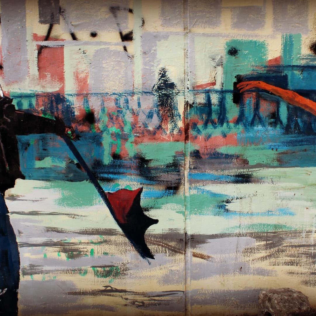 L'immagine mostra un murales che raffigura una scena dinamica e contrastante tra due figure. A sinistra, c'è una persona vestita con abiti scuri e un cappuccio, che tiene un ombrello mentre sembra quasi essere in movimento, con una gamba sollevata come se stesse per fare un passo danzante. A destra, c'è una ballerina vestita in un abito chiaro, probabilmente una tutù da danza classica, che si trova in una posa elegante con una gamba alzata all'indietro e le braccia aperte. Lo sfondo è astratto, con colori vivaci e pennellate ampie che suggeriscono edifici e una scena urbana. Il contrasto tra lo stile di abbigliamento scuro e casual del primo personaggio e la grazia e la leggerezza della ballerina crea un'immagine ricca di movimento e significato.