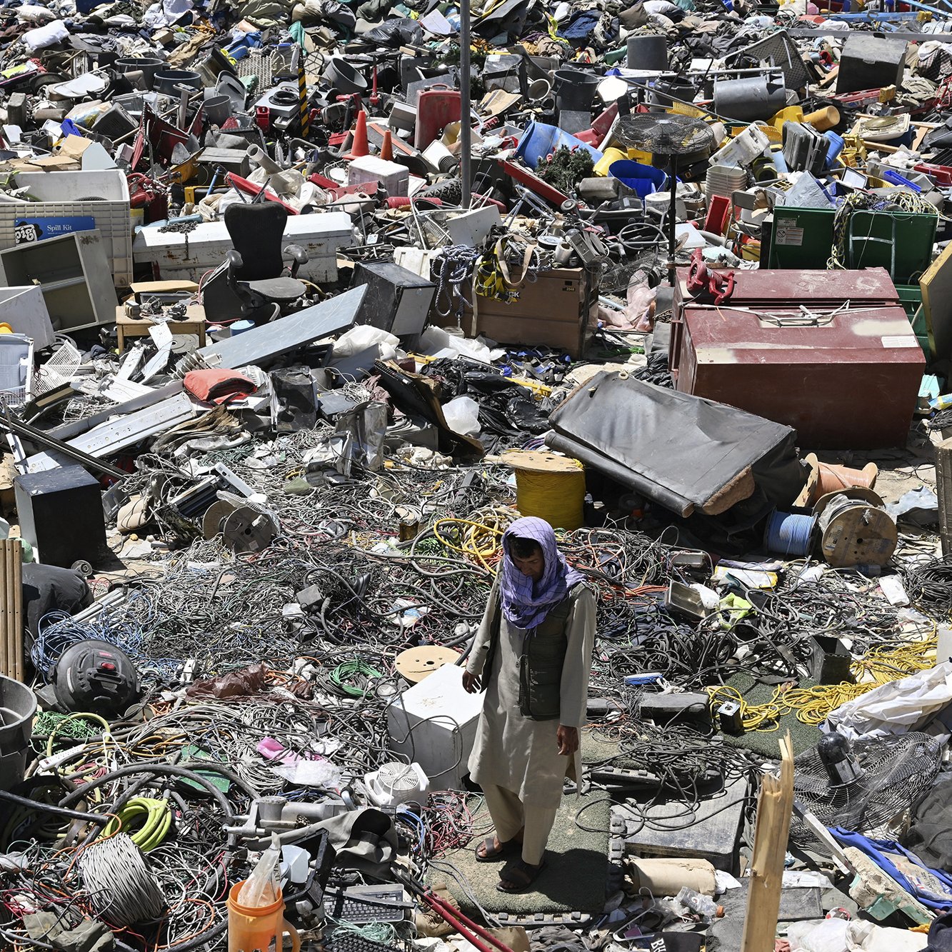 L'image montre un vaste amoncellement de déchets et de débris dans un espace encombré. On y voit des objets divers tels que des appareils électroménagers, des câbles, des pneus, et d'autres débris. Il y a aussi une personne qui marche au milieu de ce désordre, donnant une idée de l'échelle des détritus présents. L'environnement semble désordonné et négligé, illustrant une scène de récupération ou de désindustrialisation.