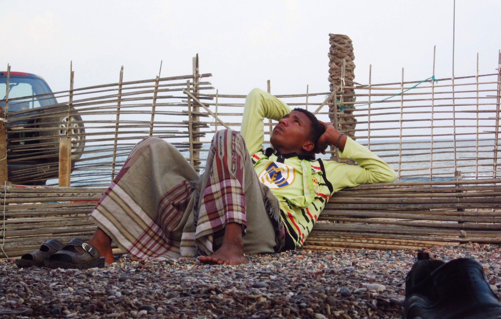Un garçon allongé, pensif, contre une barrière en bois, sur une plage de gravier.