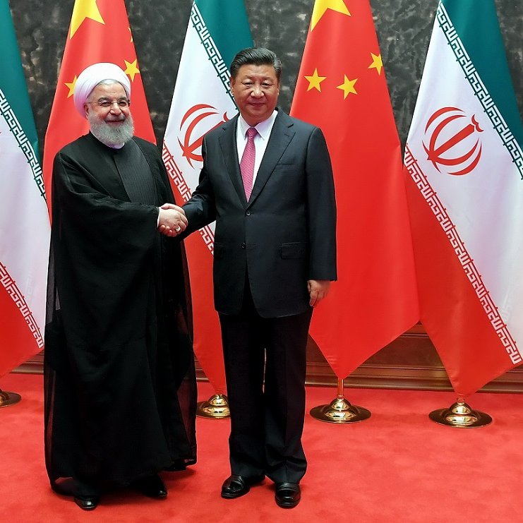 The image depicts two men shaking hands, standing in front of flags of Iran and China. The man on the left is wearing a black robe and white turban, representing Iran, while the man on the right is dressed in a suit. They appear to be in a formal setting, likely symbolizing a diplomatic meeting between the two countries. The background features a red carpet and prominently displayed national flags.