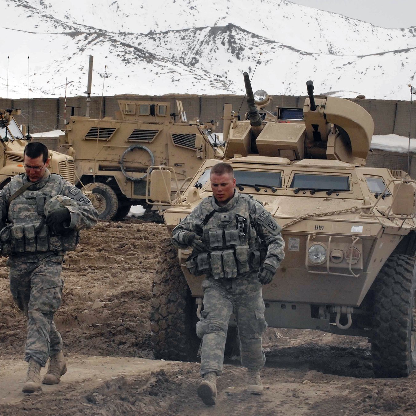 L'image montre deux soldats marchant dans un environnement rocailleux, probablement dans une zone militaire. Ils portent des uniformes camouflage et des gilets tactiques. En arrière-plan, on peut voir des véhicules blindés semblant robustes, probablement utilisés pour des opérations militaires, avec des montagnes enneigées en toile de fond. L'atmosphère semble sérieuse et empreinte d'une certaine rigueur militaire.