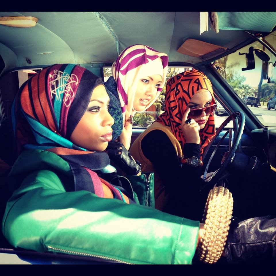 L'image montre trois femmes à l'intérieur d'une voiture. Elles portent des hijabs colorés avec des motifs variés. L'une d'elles est au volant, tandis que les deux autres semblent attentives à ce qui se passe à l'extérieur. L'ambiance est dynamique et moderne, mettant en valeur des styles vestimentaires tendance. Les couleurs vives des hijabs contrastent avec l'intérieur de la voiture, créant une atmosphère vibrante.