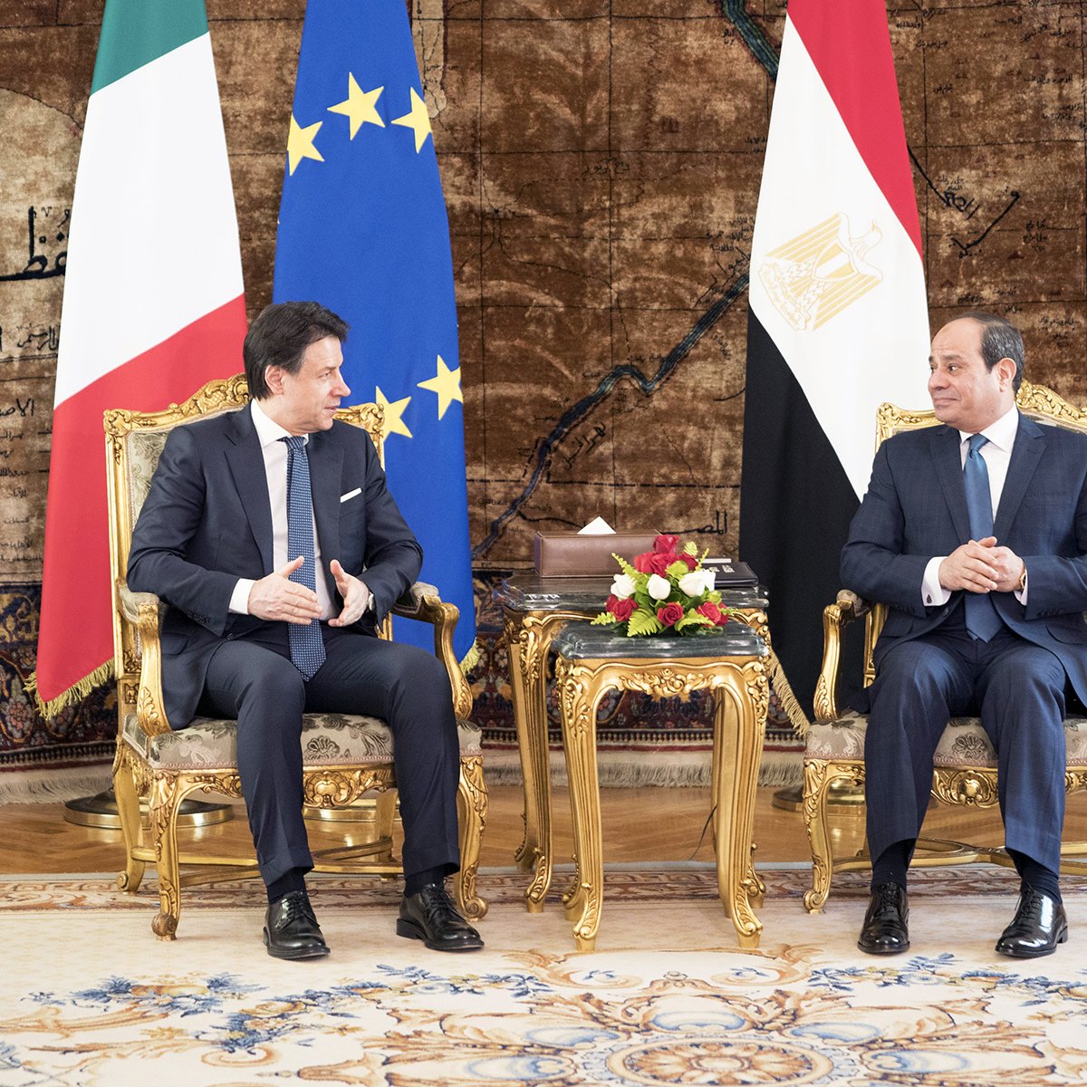 L'image montre deux hommes assis dans des fauteuils dorés dans un cadre élégant. À gauche, un homme en costume noir discute avec un homme en costume bleu à droite. Derrière eux, on peut voir des drapeaux italien et égyptien, ainsi qu'un drapeau de l'Union européenne. Le sol est recouvert d'un tapis richement décoré, et un arrangement floral est placé sur la table entre eux. L'atmosphère semble formelle, suggérant une réunion diplomatique ou une conversation importante.
