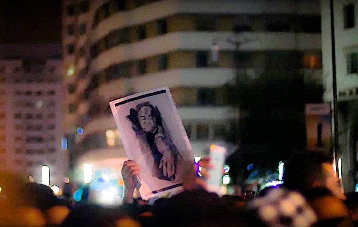 L'image montre une foule rassemblée lors d'une manifestation nocturne. Parmi les participants, une personne tient une grande affiche illustrant un portrait. Le fond de la scène est entouré de bâtiments éclairés, contribuant à l'atmosphère vibrante de l'événement. La photo semble capturer un moment important d'expression collective et de solidarité.