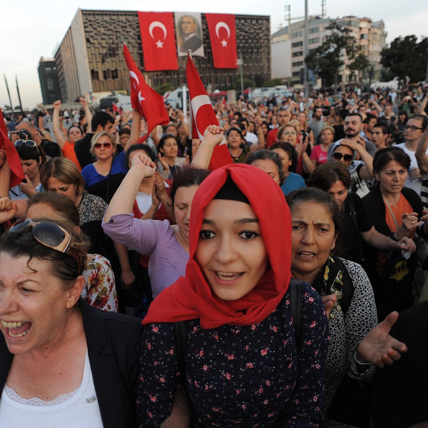 L'image montre une grande foule de personnes rassemblées, probablement lors d'une manifestation. Beaucoup d'entre elles tiennent des drapeaux turcs. On peut voir des expressions de passion et d'engagement sur les visages des participants. Certains semblent crier ou exprimer des émotions fortes. L'arrière-plan présente des bâtiments modernes, et il y a des affiches affichant des symboles ou des images en rapport avec la Turquie. L'atmosphère générale semble dynamique et pleine d'énergie collective.