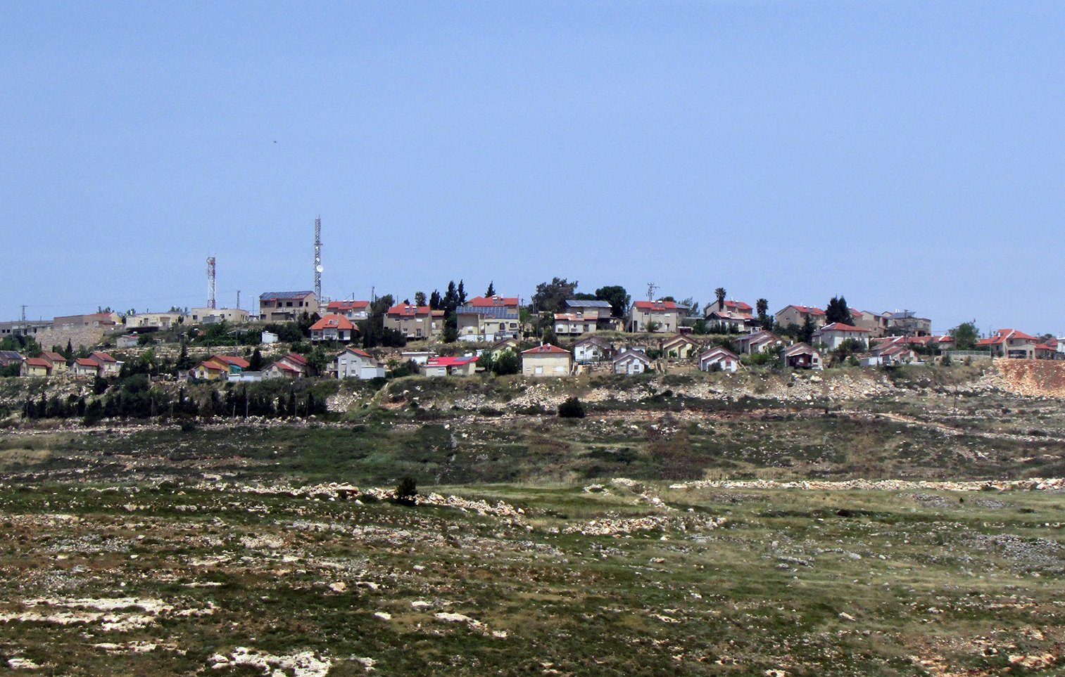 L'image montre un village situé sur une colline, avec des maisons dispersées, certaines avec des toits rouges. On peut également voir des installations comme des antennes de communication. Le paysage environnant est composé de champs et de collines vertes, suggérant une région rurale. Le ciel est clair et bleue, ce qui donne une ambiance tranquille à la scène.