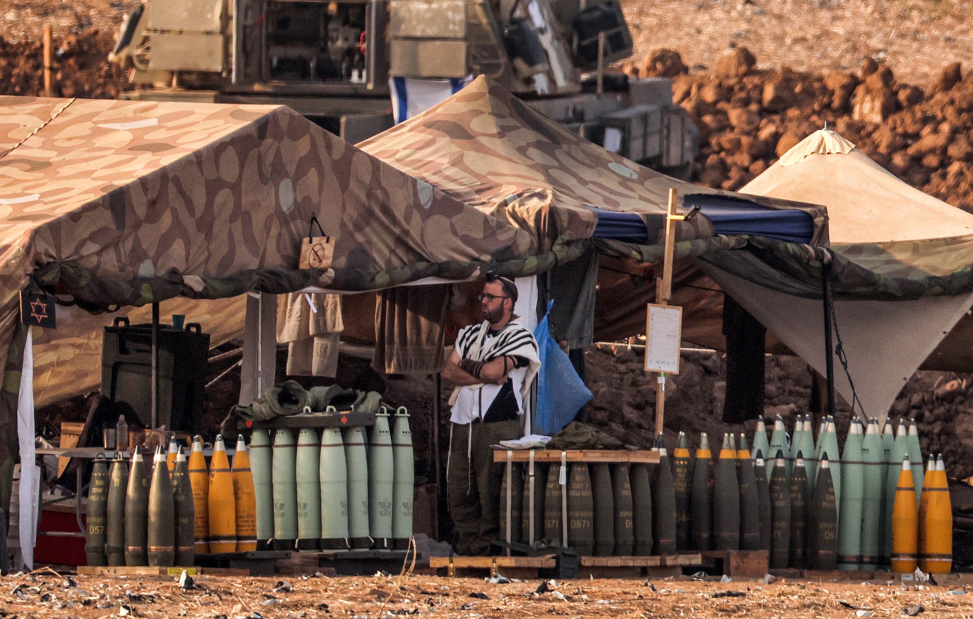 The image depicts a military encampment with tents set up in a rugged landscape. In the foreground, a soldier wearing a tallit (a traditional Jewish prayer shawl) stands near stacks of artillery shells arranged on a wooden platform. The tents are camouflaged, blending into the environment, and various military equipment is visible in the background. The setting conveys a sense of military readiness and activity.