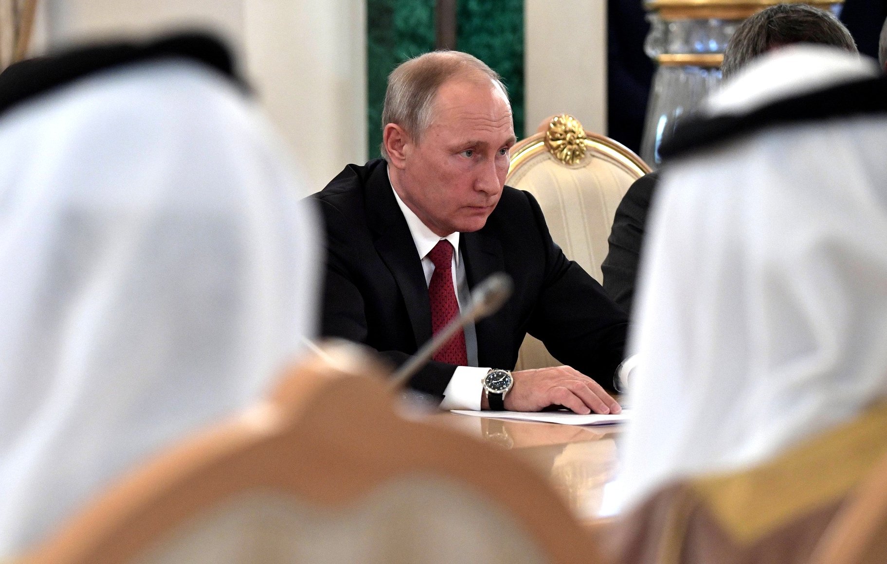 تظهر الصورة اجتماعًا رسميًا حيث يجلس رجل يرتدي بدلة ذات لون داكن. يبدو أنه مركز جداً، حيث ينظر إلى الطاولة أمامه. في الخلفية، يمكن رؤية أشخاص آخرين يرتدون الملابس التقليدية، مما يعطي انطباعًا عن حدث رسمي أو دبلوماسي يشارك فيه ممثلون من ثقافات مختلفة. يحيط بالاجتماع أجواء من الجدية والتركيز.