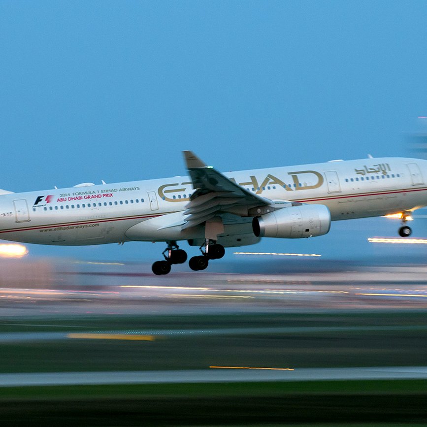 L'image montre un avion de la compagnie aérienne Etihad Airways en phase de décollage ou d'atterrissage. L'avion est partiellement flou, ce qui suggère une vitesse élevée. Le ciel apparaît dans une teinte bleue, tandis que des lumières brillantes en arrière-plan ajoutent une ambiance dynamique. L'appareil affiche la décoration et le logo caractéristiques de la compagnie.