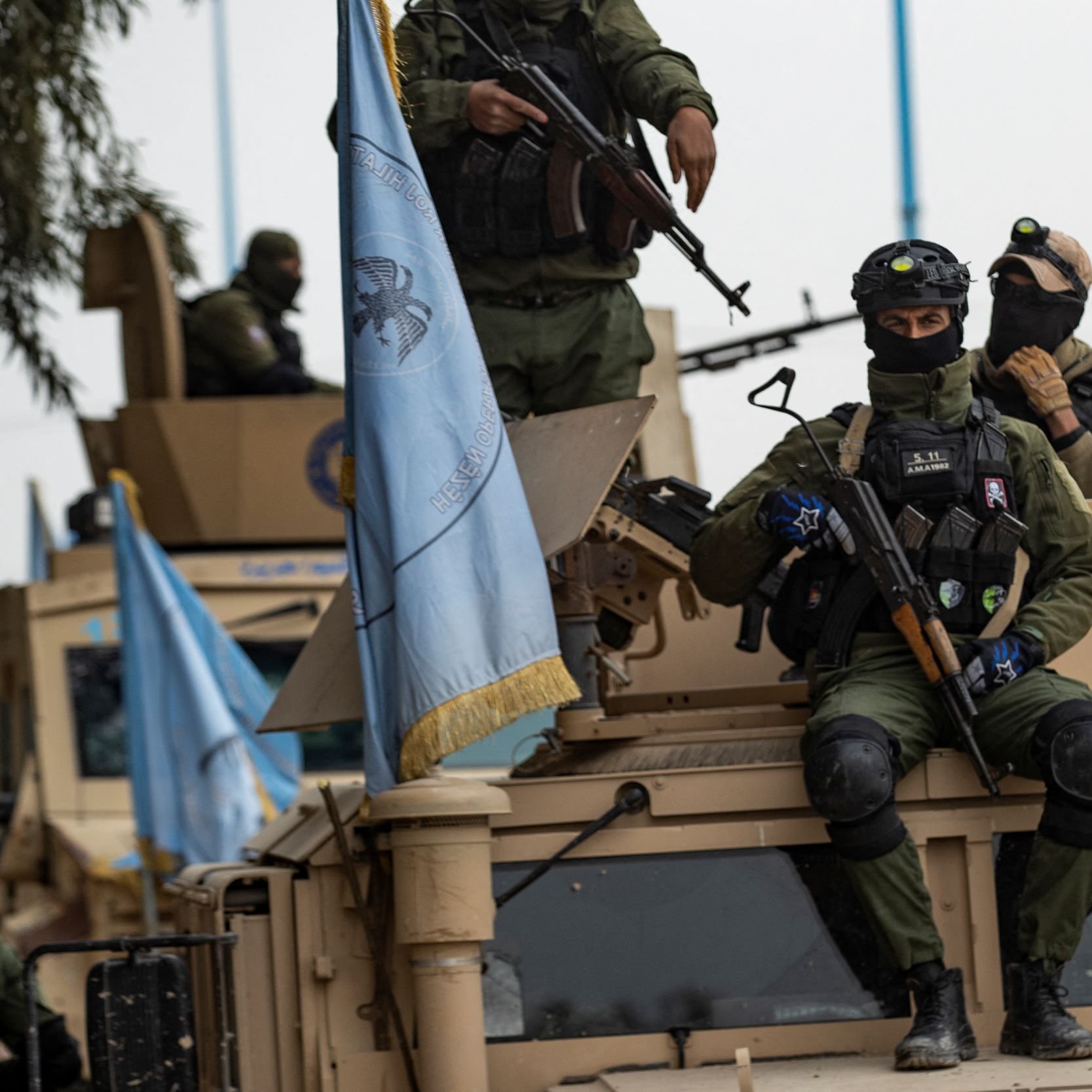 این تصویر گروهی از نیروهای مسلح را نمایش می‌دهد که روی یک وسیله نقلیه نظامی نشسته‌اند. نیروها لباس‌های نظامی تیره و کلاه‌خود دارند و برخی از آن‌ها سلاح‌های جداگانه‌ای در دست دارند. بر روی وسیله نقلیه پرچمی و نمادهای مربوط به سازمان‌های بین‌المللی یا نظامی به چشم می‌خورد. پس‌زمینه تصویر به وضوح نشان‌دهنده یک محیط خارج از شهر است.