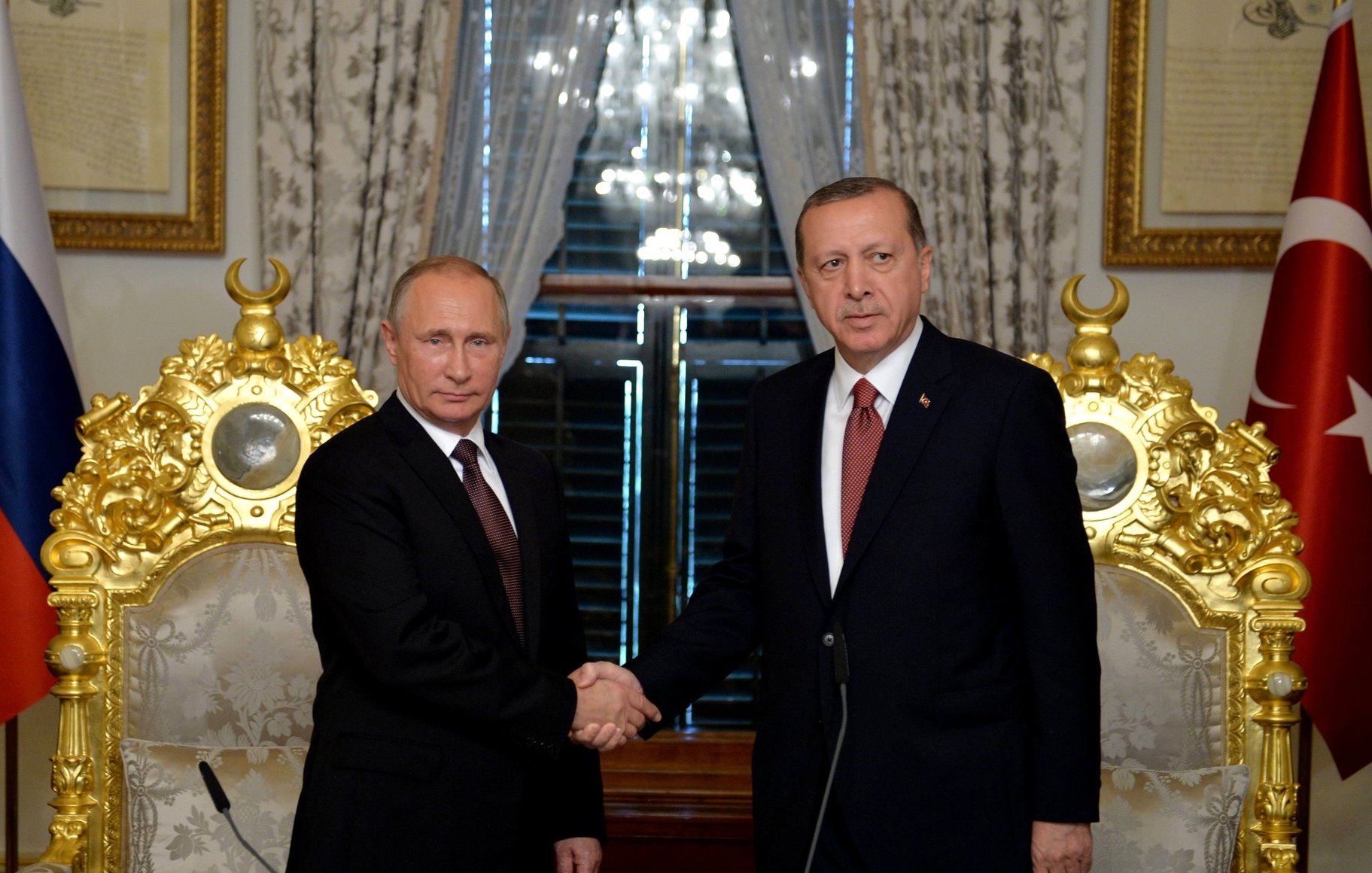 تصویر نشان‌دهنده‌ی دو نفر است که در حال握 دستی هستند. آن‌ها در یک محیط رسمی قرار دارند و در پس‌زمینه، دکوراسیون مجلل و البته دو پرچم، یکی از روسیه و دیگری از ترکیه، دیده می‌شود. این صحنه می‌تواند به یک ملاقات یا اجلاس رسمی بین مقامات دو کشور اشاره داشته باشد. فضای تصویر به طور کلی جدی و رسمی به نظر می‌رسد.