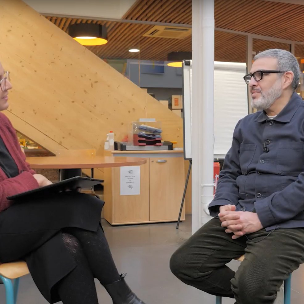 L'image montre deux personnes assises dans un espace intérieur. À gauche, une femme avec des cheveux bouclés et des lunettes, vêtue d'un pull rouge, tient une tablette. À droite, un homme portant des lunettes et une chemise bleu foncé est assis, les mains sur ses genoux. L'environnement est moderne, avec des murs en bois et des éléments de décoration qui donnent une ambiance chaleureuse. Cela semble être une interview ou une discussion.