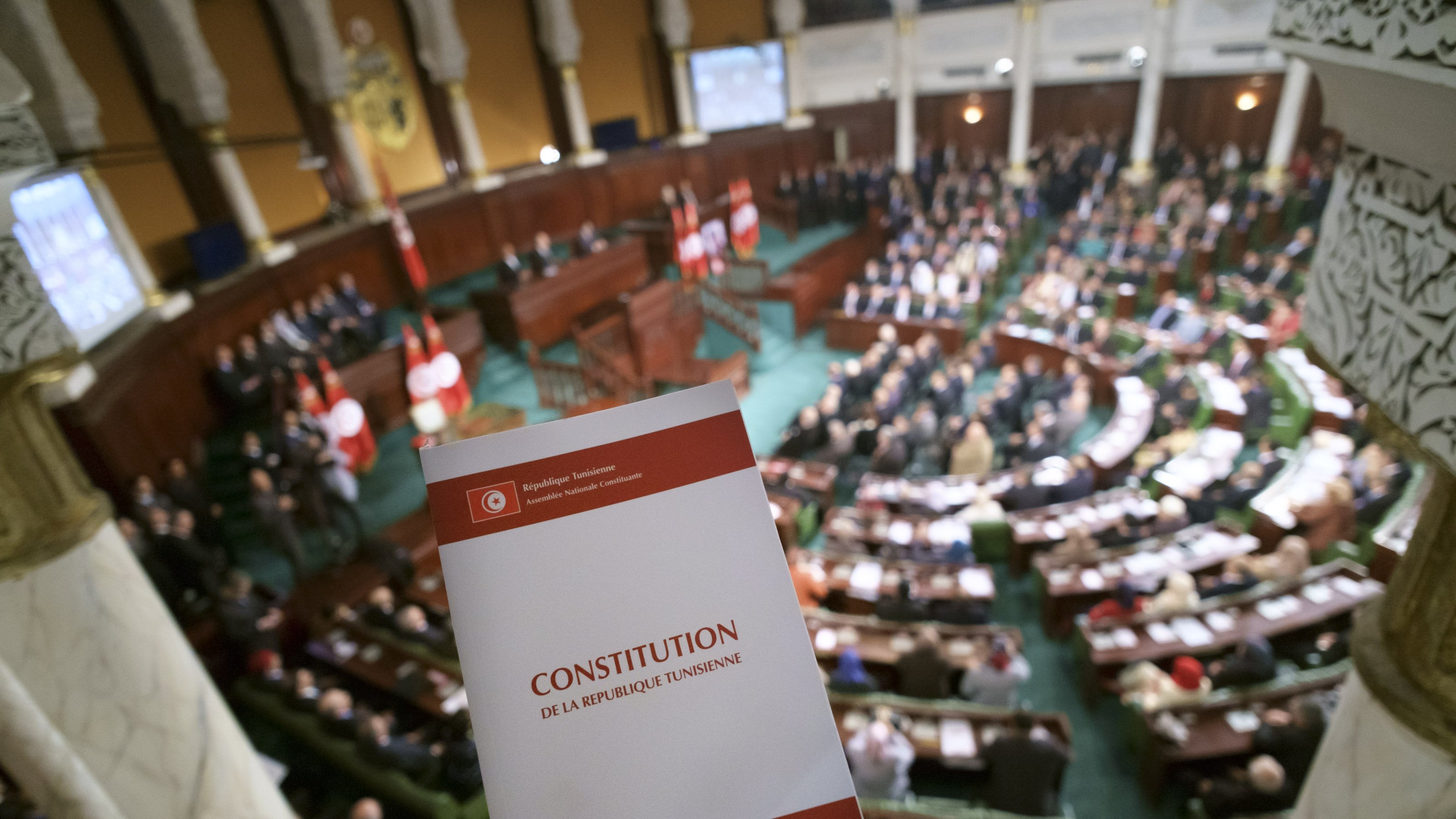 تظهر في الصورة قاعة البرلمان التونسي، حيث يوجد مجموعة كبيرة من النواب والموظفين في الجلسة. في المقدمة، هناك شخص يحمل نسخة من الدستور التونسي. الخلفية مليئة بالعلم التونسي، مما يدل على أهمية الحدث الذي يتم مناقشته في القاعة. التصميم الداخلي للقاعة يبدو أنيقًا، مع تفاصيل معمارية تقليدية.