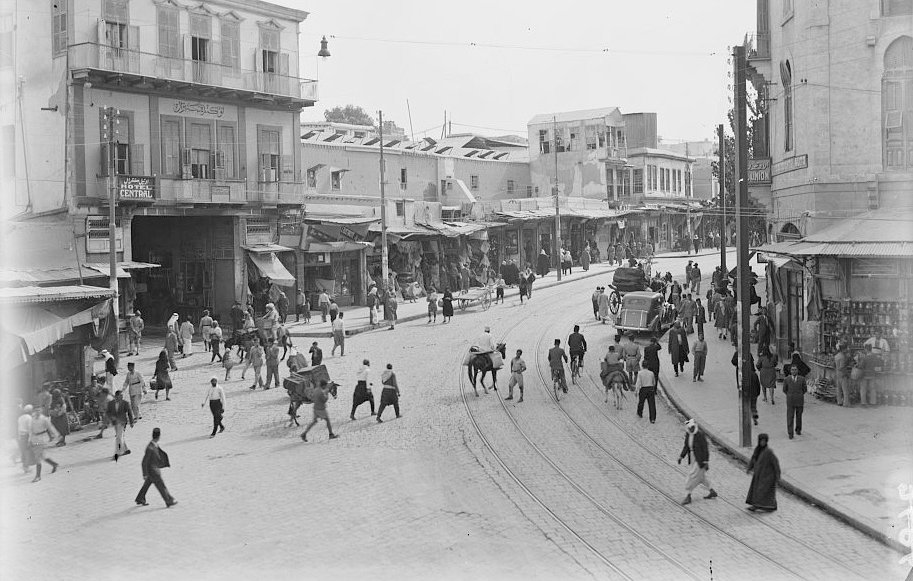 L'image montre une vue animée d'une rue commerçante dans une ville. On y voit des bâtiments à plusieurs étages avec des balcons, des étals et des commerces le long de la rue. De nombreuses personnes, y compris des piétons et des animaux, circulent. On aperçoit également une voiture ancienne sur la route. L'atmosphère semble vibrante et typique d'une époque passée, avec un mélange de textures urbaines et de la vie quotidienne.