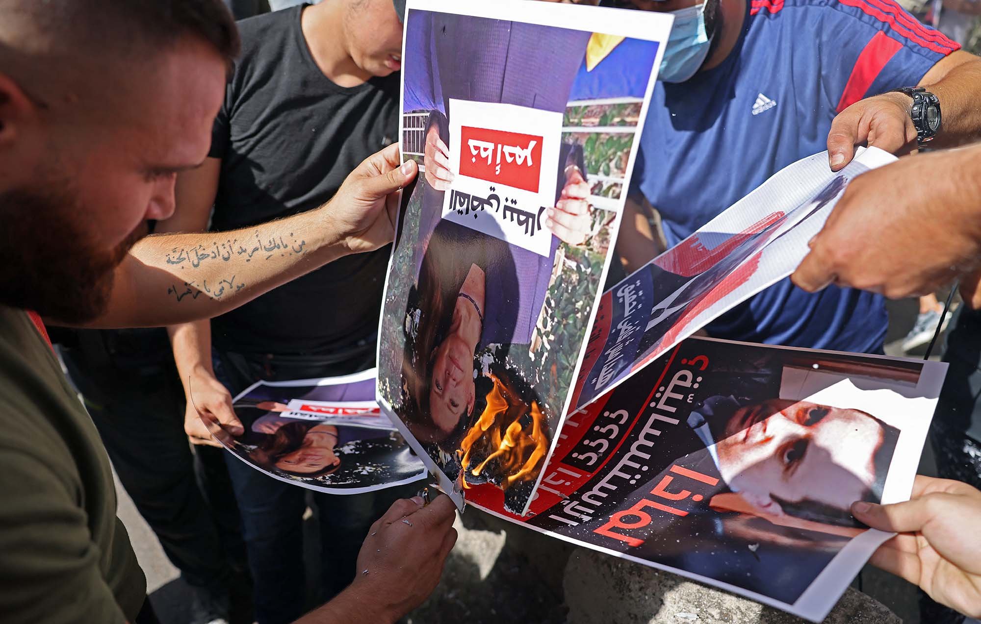 Nell'immagine si vedono diverse persone che stanno bruciando dei manifesti. I manifesti ritraggono volti, probabilmente di una figura pubblica, e sono esposti a una fiamma. Le persone sembrano partecipare attivamente a questa protesta o atto simbolico, esprimendo il loro discontento. L'atmosfera è di intensa emozione e opposizione.