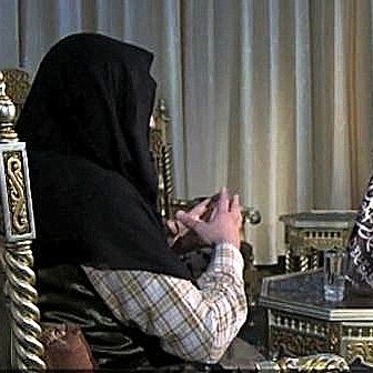 L'image montre deux personnes assises dans un cadre décoratif. L'un des individus porte un hijab et est assis sur un fauteuil orné, tandis que l'autre, en costume, est également assis sur un fauteuil face à lui, tenant un livre. L'environnement semble richement décoré avec des motifs orientaux et des lumières suspendues. L'atmosphère de la scène semble être celle d'une conversation sérieuse.
