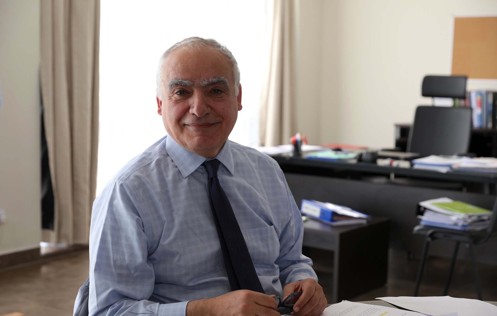 L'immagine mostra un uomo sorridente seduto a un tavolo in un ambiente di lavoro. Indossa una camicia e una cravatta, e sembra essere concentrato su alcune carte e documenti posti davanti a lui. Sullo sfondo, si intravedono elementi di un ufficio, come una scrivania con vari materiali e un ambiente luminoso grazie alla luce naturale che entra dalla finestra.