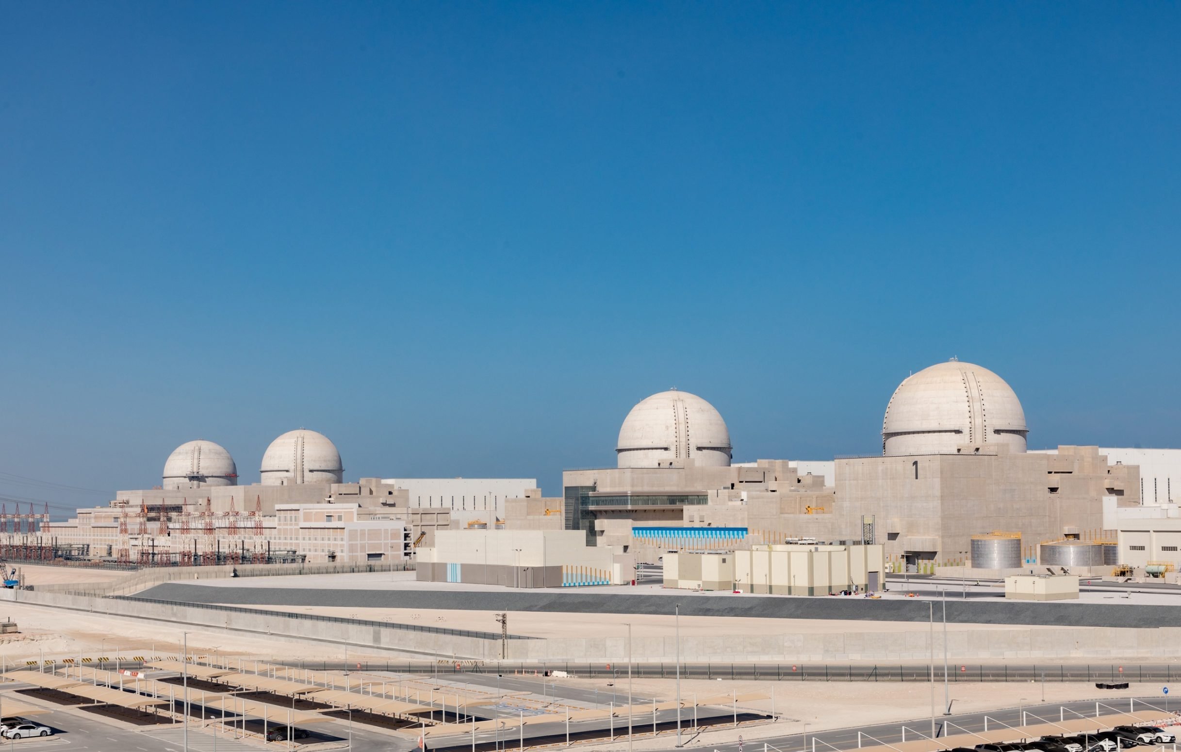 L'immagine mostra una centrale nucleare moderna, caratterizzata da tre imponenti cupole bianche che emergono dal complesso. La struttura presenta un design industriale, con vari edifici di colore chiaro che si estendono in un'area ampia e ben organizzata. Il cielo è sereno e blu, suggerendo una bella giornata. In primo piano, si può notare un'area ben curata e delle strade di accesso, tipiche delle installazioni di questo tipo.