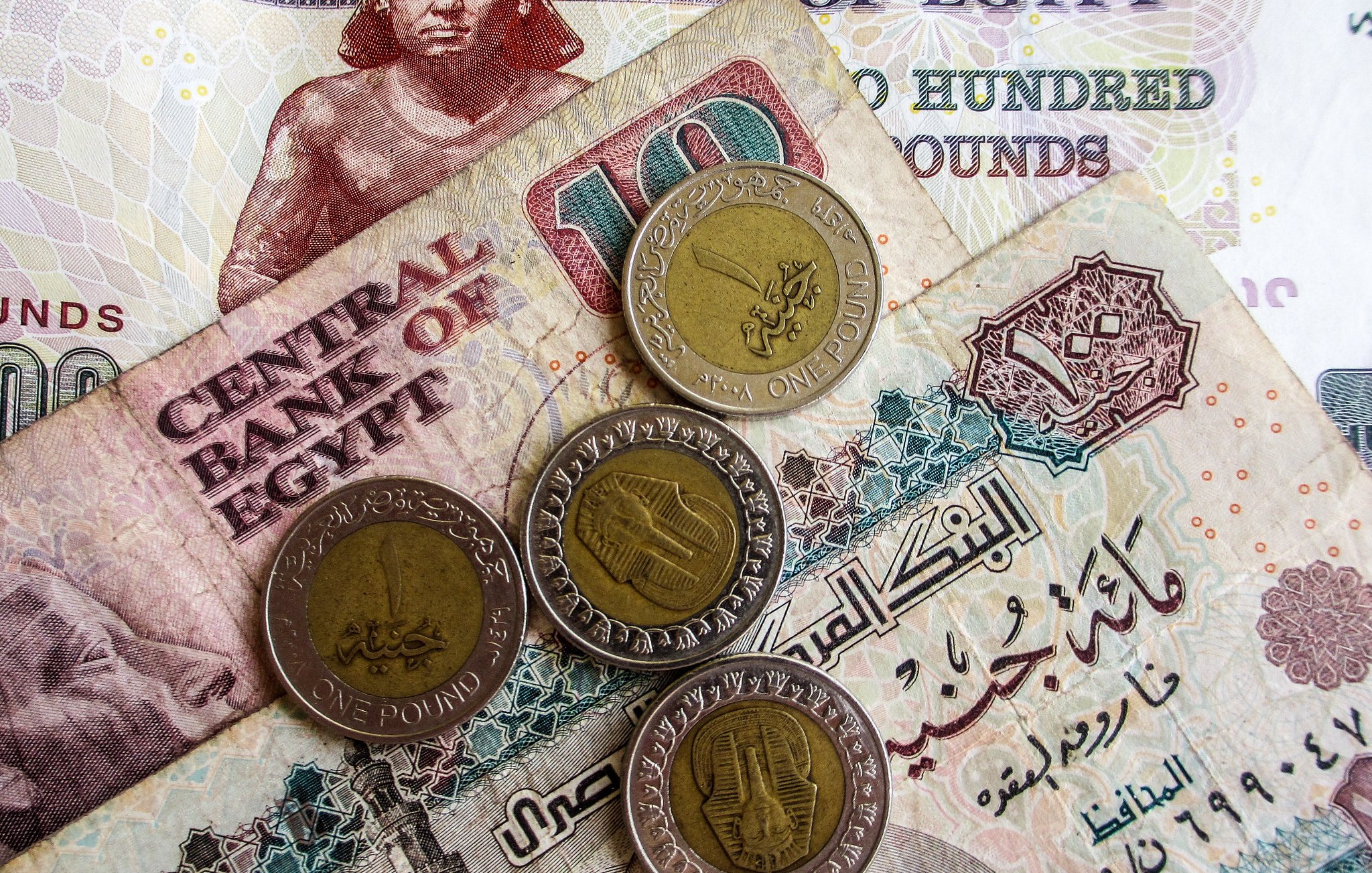 تظهر في الصورة مجموعة من العملات المصرية. هناك ورقة نقدية من فئة المائة جنيه بالإضافة إلى فئة العشرة جنيه، مع عدد من العملات المعدنية من فئة الجنيه الواحد. التصميمات المختلفة توضح رموز ومعالم ثقافية مرتبطة بمصر، مما يعكس التنوع في العملة المصرية.