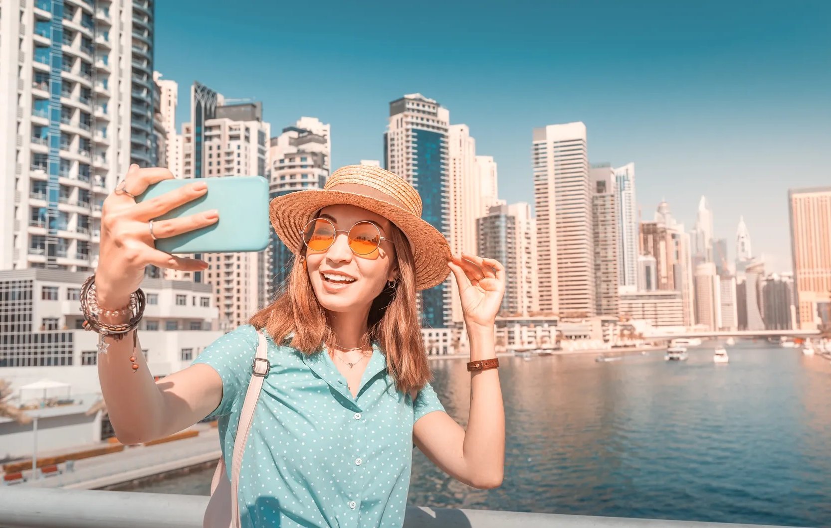 L'image montre une jeune femme prenant un selfie au bord de l'eau, probablement dans une ville dynamique. Elle porte un chapeau en paille et des lunettes de soleil orange, ajoutant une touche estivale à son look. Elle sourit joyeusement, semblant profiter de son moment. En arrière-plan, on peut voir des gratte-ciel modernes et un port avec des bateaux, évoquant un environnement urbain animé et ensoleillé.