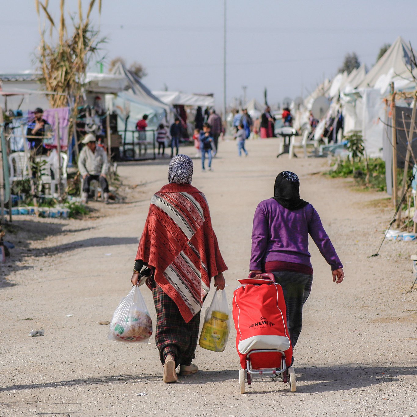 L'image montre un camp de réfugiés ou un espace similaire, avec plusieurs tentes dressées de chaque côté d'un chemin central. Deux femmes marchent le long de ce chemin, portant des sacs et un chariot. L'une d'elles porte une couverture ou un châle coloré. En arrière-plan, on peut apercevoir d'autres personnes et des tentes, suggérant une atmosphère de communauté. Le paysage semble rural et ensoleillé, avec des éléments de végétation autour.