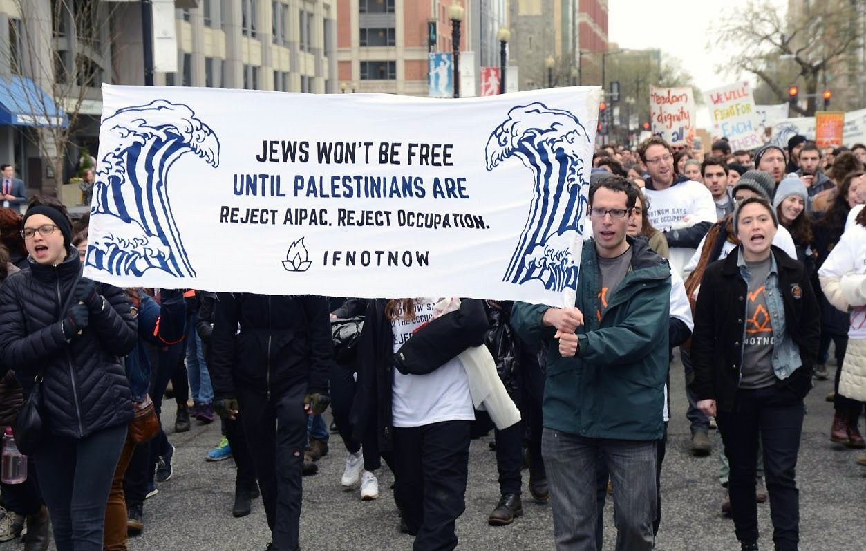 The image shows a group of protesters marching together, holding a large banner that reads, "Jews won't be free until Palestinians are. Reject AIPAC. Reject occupation." The banner features artistic waves, and the participants appear to be actively engaged in the demonstration, dressed in casual winter clothing. The background includes urban buildings and other protesters. The atmosphere conveys a sense of solidarity and activism focused on social justice issues.