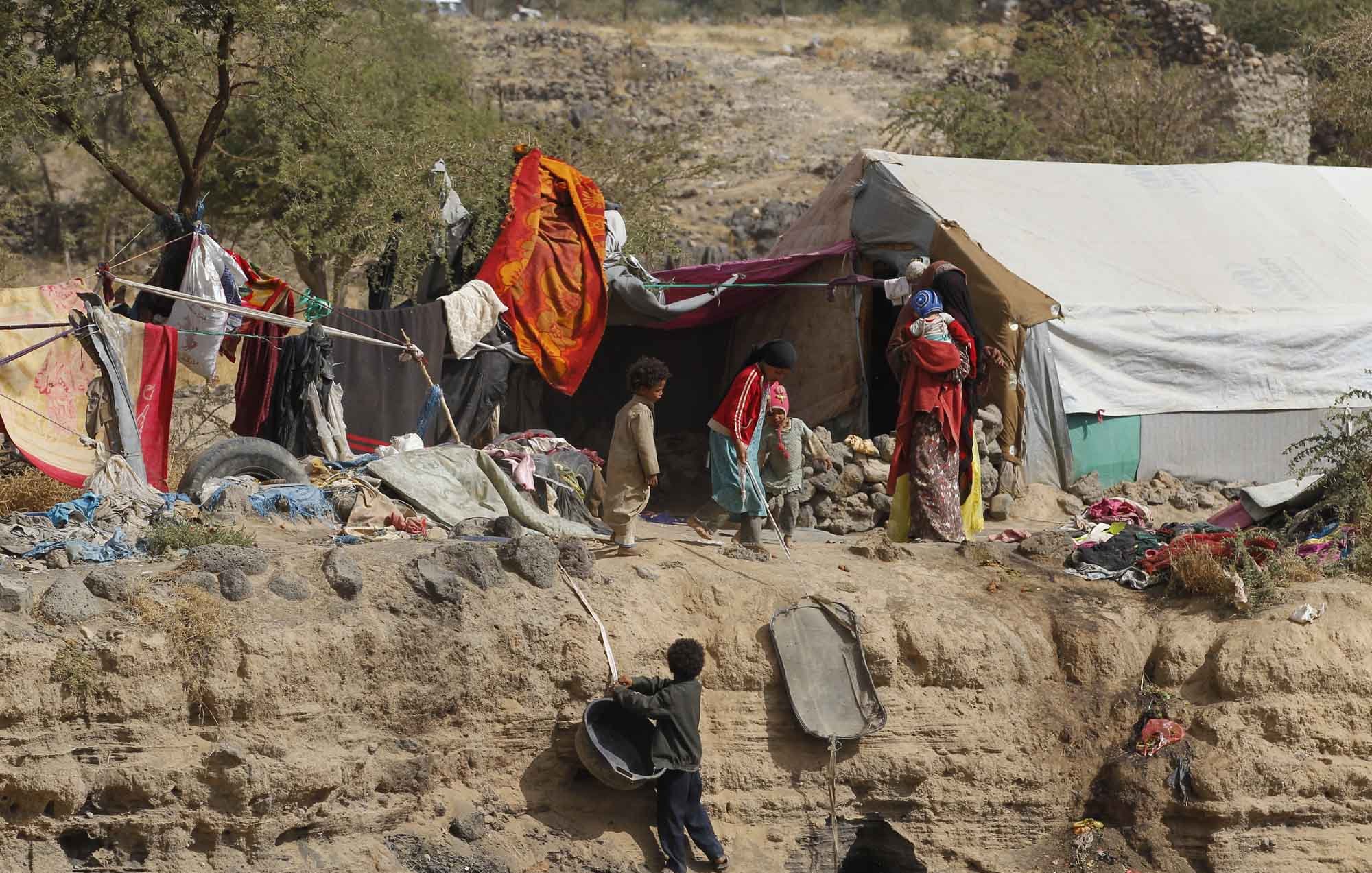 L'image montre un paysage difficile, probablement dans une région aride. On y voit des habitations précaires constituées de bâches et de morceaux de tissu, témoignant de conditions de vie modestes. Quelques enfants jouent près des abris, tandis que d'autres semblent s'affairer. L'environnement est marqué par des terrains rocheux et des éléments naturels, suggérant une situation de vulnérabilité. La scène évoque des thèmes liés à la pauvreté et à la résilience des communautés vivant dans des conditions précaires.