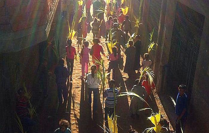 الصورة تظهر مجموعة من الأشخاص في شارع ضيق، يجتمعون معًا وهم يحملون أوراق نباتية طويلة، ربما يمثلون احتفالًا أو مهرجانًا. الأفراد يرتدون ملابس ملونة ويبدون متحمسين. تضيء الشمس المكان، مما يضيف جواً مميزاً للصورة، ويعطي انطباعًا بالحيوية والفرح خلال الفعالية.