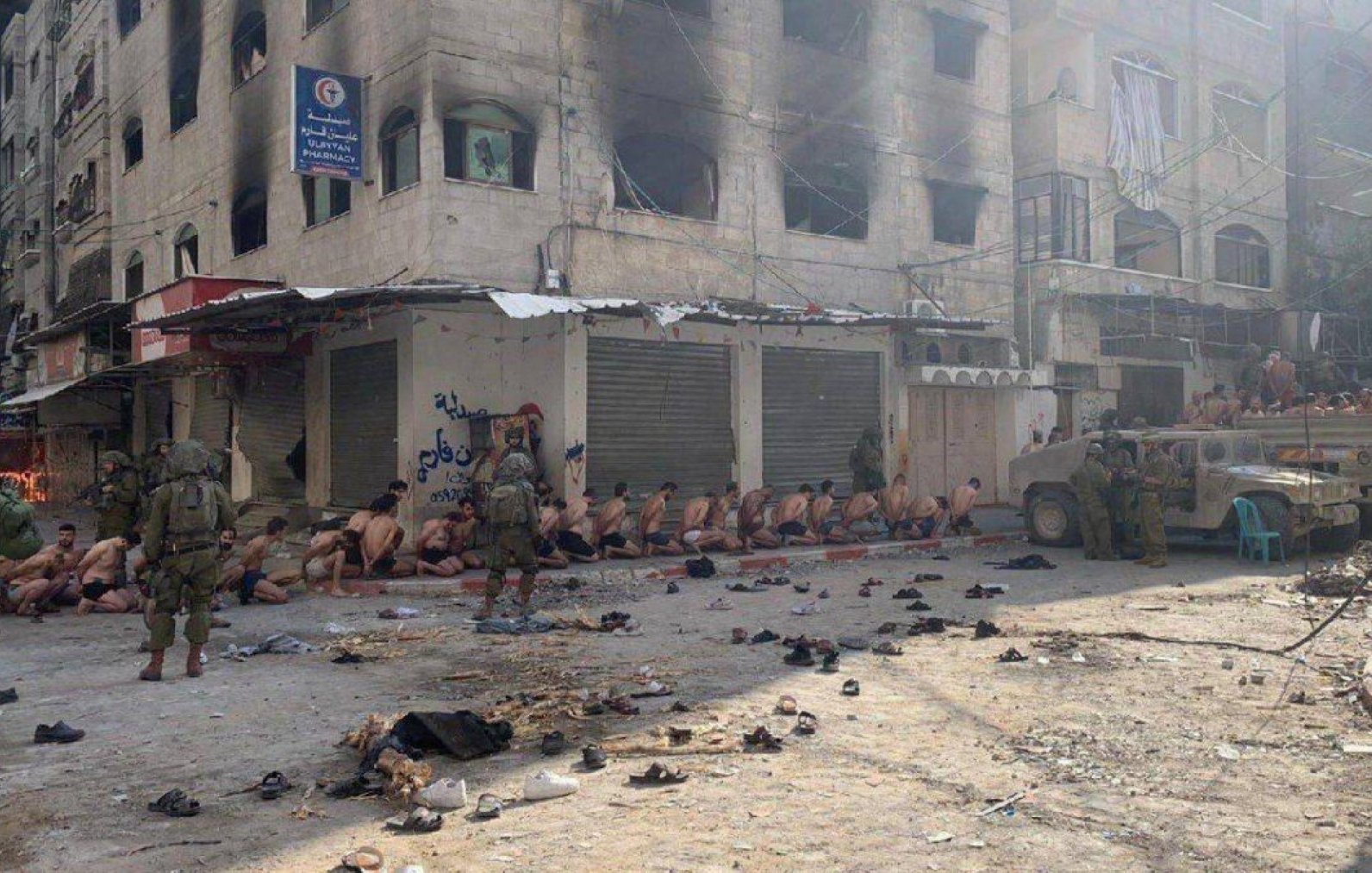 Immagine di un gruppo di persone su un marciapiede, sotto sorveglianza militare, in un contesto urbano.