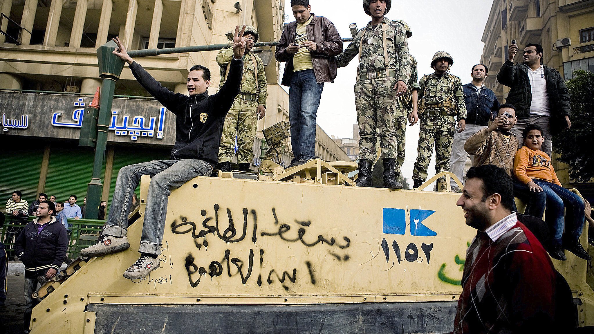 L'image montre un groupe de personnes rassemblées autour d'un véhicule militaire dans une ville. On peut voir des soldats en uniforme sur le véhicule, tandis que d'autres personnes, probablement des civils, interagissent avec eux. L'atmosphère semble tendue, mais aussi pleine d'énergie, comme si une sorte de manifestation ou de rassemblement avait lieu. L'arrière-plan montre des bâtiments urbains et des enseignes en arabe, ce qui suggère que la scène se déroule dans un pays du Moyen-Orient.