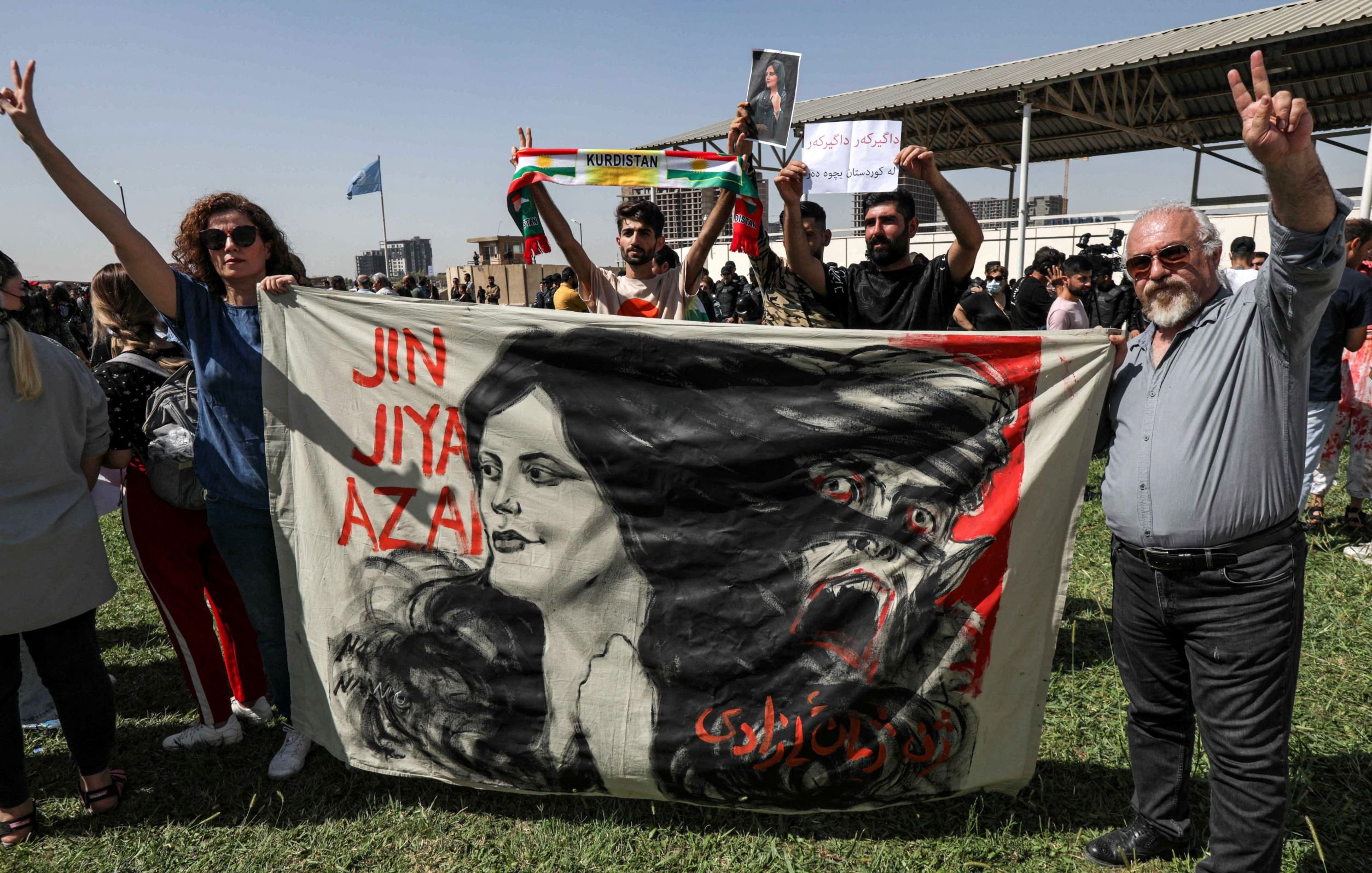 در این تصویر، افراد مختلفی در یک تجمع یا تظاهرات حضور دارند. دو نفر در جلو در حال نمایش یک بنر بزرگ هستند که تصویر زنی با نام "زن، زندگی، آزادی" و یک سر حیوان را نشان می‌دهد. در پس‌زمینه، عده‌ای دیگر در حال برگزاری تظاهرات هستند و پلاکاردهایی در دست دارند. فضای تصویر نشان‌دهنده همبستگی و جنبش‌های اجتماعی است. افراد در حال نشان دادن علامت پیروزی هستند و احساسات قوی را ابراز می‌کنند.