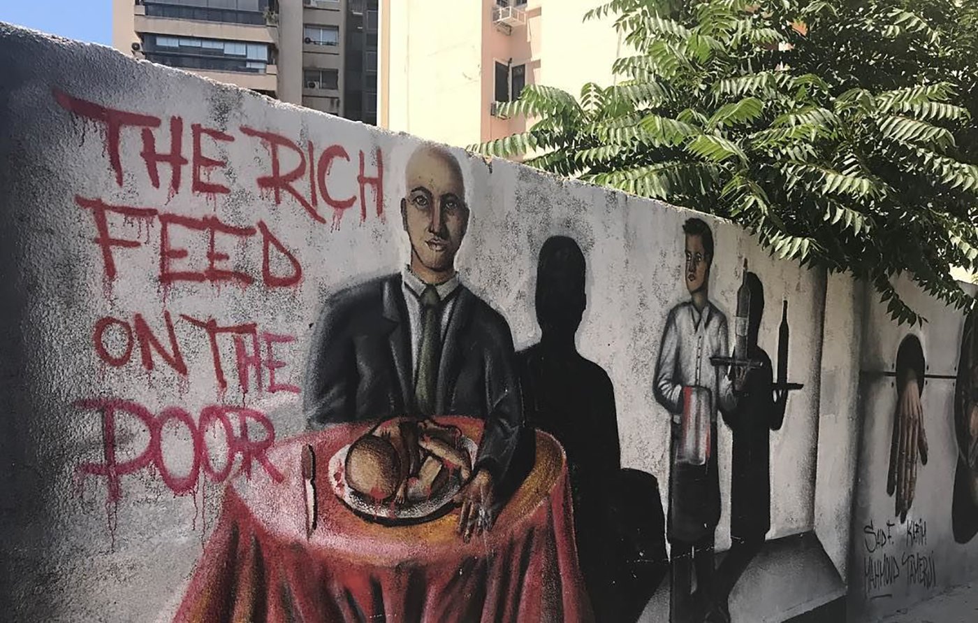 تظهر في الصورة جداريات على حائط، حيث يبرز جزء من العمل الفني رسما لشخص يجلس على طاولة، مرتديًا بدلة، ويبدو أنه يتناول الطعام. خلفه توجد ظلال لشخصيات أخرى، بما في ذلك شخص يرتدي ملابس عامل، مما يعكس تباينًا بين الطبقات الاجتماعية. تزين الجدارية أيضًا عبارة باللغة الإنجليزية "THE RICH FEED ON THE POOR" (الأغنياء يتغذون على الفقراء)، مما يوحي برسالة اجتماعية قوية حول الفوارق الطبقية والاستغلال. في الخلفية، يمكن رؤية أشجار وبعض المباني السكنية، مما يشير إلى أن هذه الجدارية تقع في منطقة حضرية.