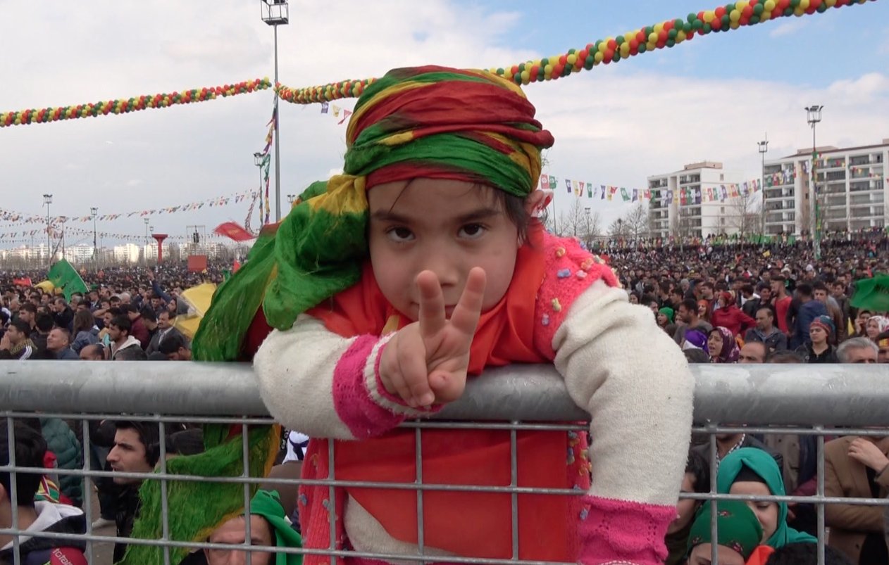 L'image montre une jeune fille participant à un grand rassemblement. Elle est vêtue de vêtements colorés, portant un foulard aux teintes vives. La fillette fait un signe de paix avec ses doigts, pendant qu'elle se penche sur une barrière. En arrière-plan, une grande foule est présente, témoignant d'un événement festif ou d'une manifestation populaire, avec des décorations colorées suspendues au-dessus. L'ambiance semble joyeuse et engagée.