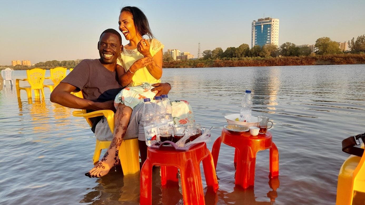 L'image montre un couple assis sur des chaises jaunes au bord de l'eau. Ils semblent heureux et détendus, avec un grand bâtiment en arrière-plan. Des bouteilles d'eau et des tasses sont posées sur des petites tables à proximité. Le cadre est serein, avec un ciel dégagé et l'eau qui reflète la lumière du soleil, créant une ambiance chaleureuse.