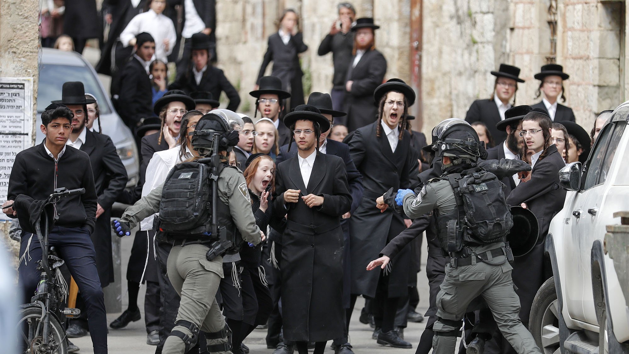 The image depicts a tense confrontation between a group of individuals dressed in traditional Orthodox Jewish attire and armed security personnel. The group appears to be expressing strong emotions, possibly protest or unrest, while the security officers, equipped with tactical gear, seem to be engaging with them. The setting appears to be an urban area, with a mixture of traditional and modern elements visible in the background, including vehicles and stone buildings. The scene captures a moment of conflict or significant social interaction.