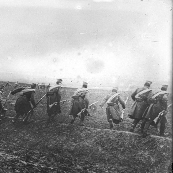 L'image montre un groupe de soldats marchant en formation sur un terrain accidenté. Ils portent des uniformes militaires, avec des chapeaux distinctifs, et semblent se déplacer dans une direction déterminée. Le paysage est marqué par un ciel nuageux et une ambiance sombre, suggérant peut-être un contexte de guerre ou de mouvement stratégique dans un environnement rural. Le sol est boueux, ce qui rend leur avance difficile. L'image est en noir et blanc, renforçant le caractère historique et poignant de la scène.